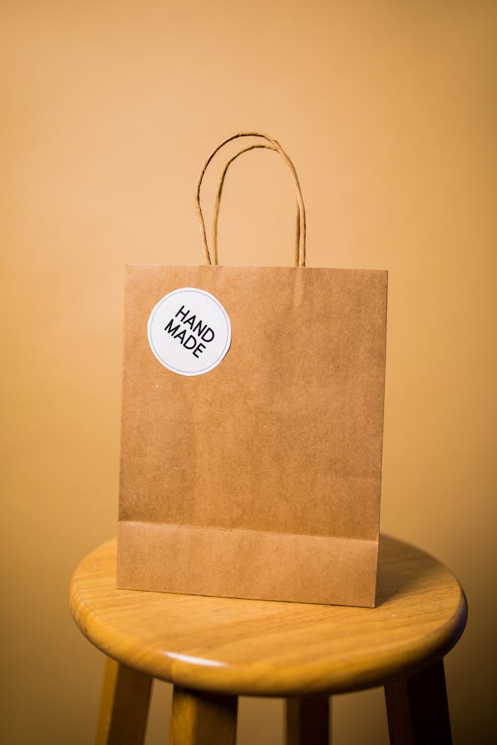 Simple brown paper bag labeled 'hand made' on a wooden stool against a beige background.